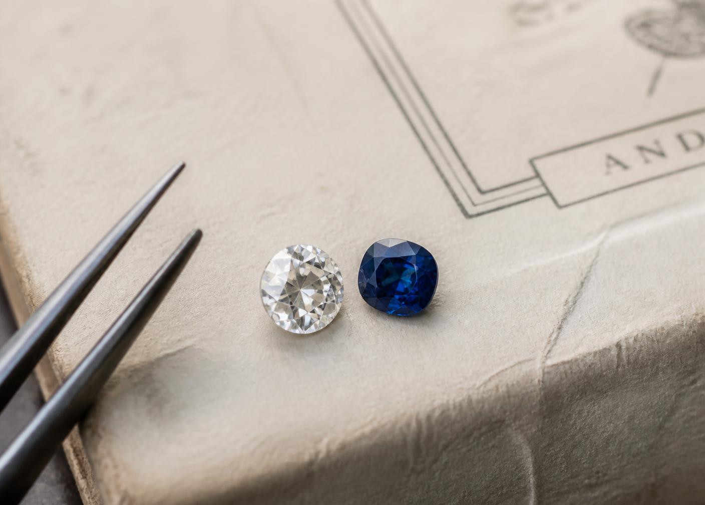 A loose diamond and sapphire resting on a paper gemstone parcel beside tweezers